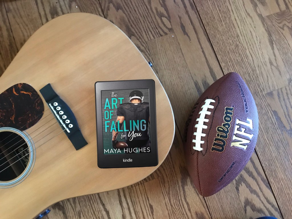 A kindle with the cover art of "The Art of Falling for You" on top of an acoustic guitar, with a football on the hardwood floor next to it.